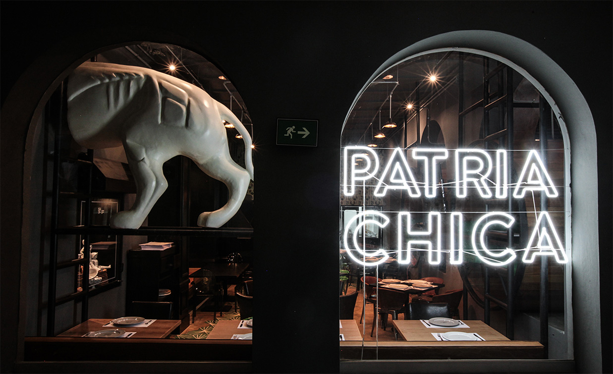 Fachada exterior del restaurante Patria Chica con letrero iluminado y dinosaurio decorativo, construcción comercial Fer Et Ciment León